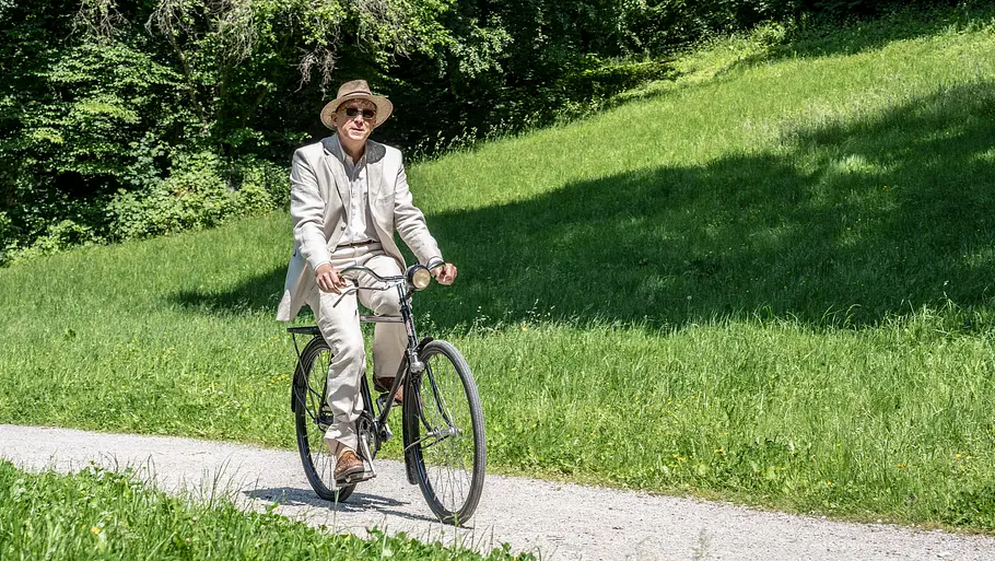 Die Ferien des Monsieur Murot Ermittler Murot fährt mit einem Fahrrad an einer grünen Wiese vorbei.
