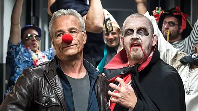 Klaus J. Behrendt (l.) steht mit Clowsnase neben Dietmar Bär, der ein Vampirkostüm trägt. Hinter ihnen sind fröhliche Personen in Kostümen. - Foto: WDR/Thomas Kost