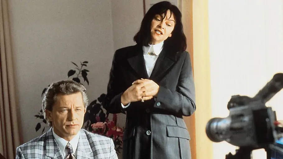 Miroslav Nemec sitzt in einem Anzug vor der Kamera. Hinter ihm steht eine Frau in einem schwarzen Fit.