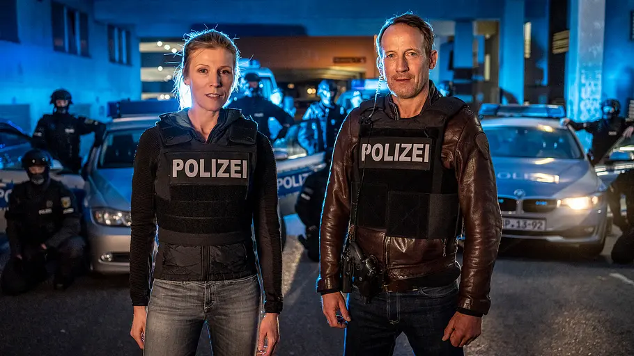 Grosz und Falke stehen in Polizeimontur vor Polizeiwagen.
