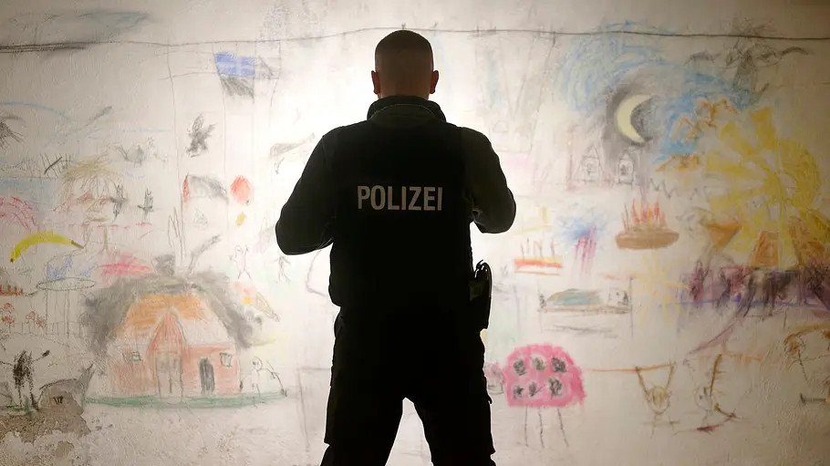 Edin Hasanovic steht mit dem Rücken zur Kamera vor einer Wand, die überseht mir KInderzeichnungen ist und schaut sich die Bilder mit einer Taschenlampe an.