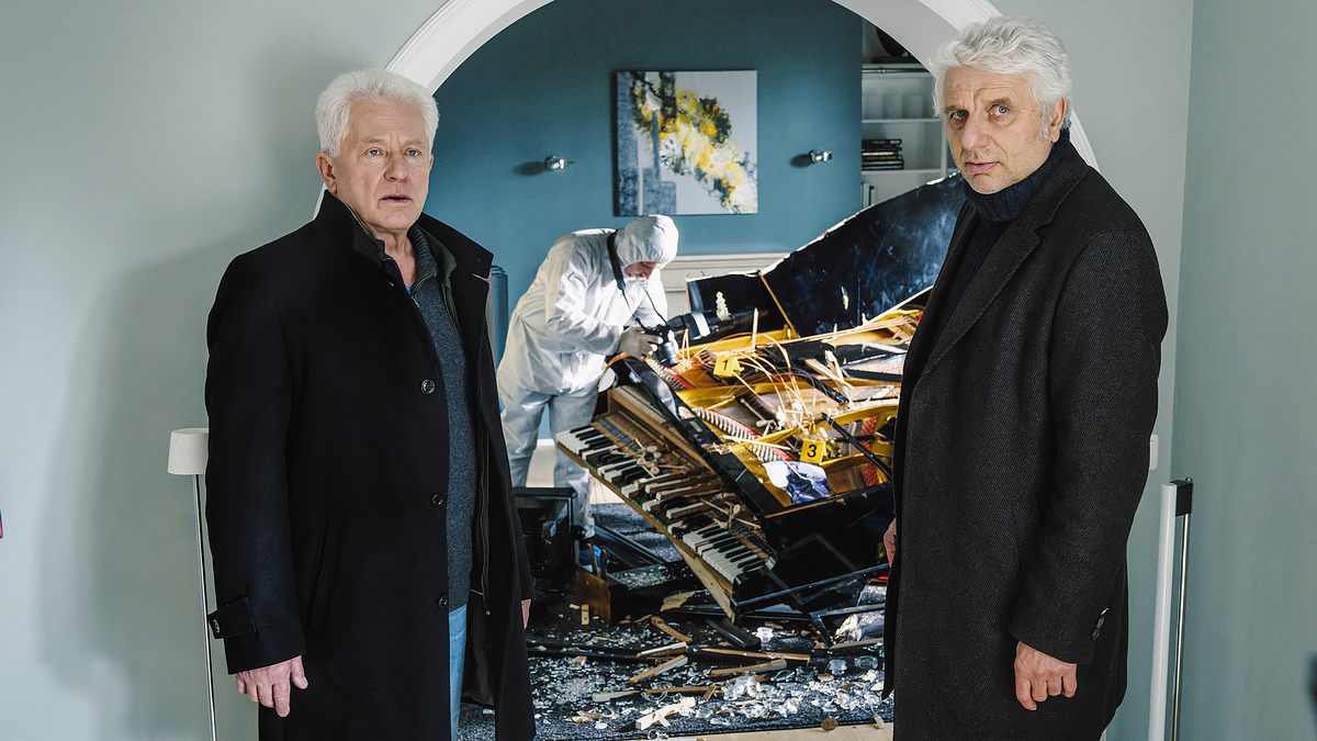 Tatort München Udo Wachtveitl (links) und Miroslav Nemec stehen in einem Wohnhaus. Beide tragen schwarze Mäntel. Hinter ihnen ist eine Person der Spurensicherung, die ein zertrümmertes Klavier inspiziert.