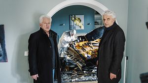 Udo Wachtveitl (links) und Miroslav Nemec stehen in einem Wohnhaus. Beide tragen schwarze Mäntel. Hinter ihnen ist eine Person der Spurensicherung, die ein zertrümmertes Klavier inspiziert.   - Foto: BR/NEUESUPER GmbH/Hendrik Heiden