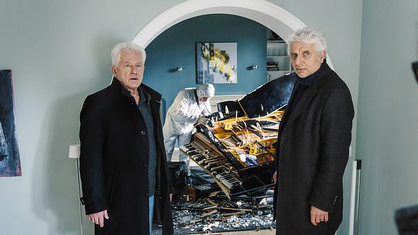 Udo Wachtveitl (links) und Miroslav Nemec stehen in einem Wohnhaus. Beide tragen schwarze Mäntel. Hinter ihnen ist eine Person der Spurensicherung, die ein zertrümmertes Klavier inspiziert.   - Foto: BR/NEUESUPER GmbH/Hendrik Heiden