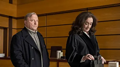 Axel Prahl und Mechthild Großmann stehen für den Münsteraner „Tatort“ in einem Gerichtssaal. - Foto: MDR/WDR/Martin Valentin Menke