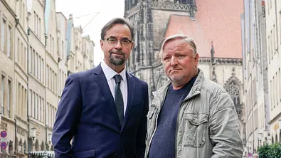 Jan Josef Liefers (l.) steht im Anzug neben einem legere gekleideten Axel Prahl in der Innenstadt Münsters. - Foto: WDR/Bavaria Fiction GmbH/Thomas Kost
