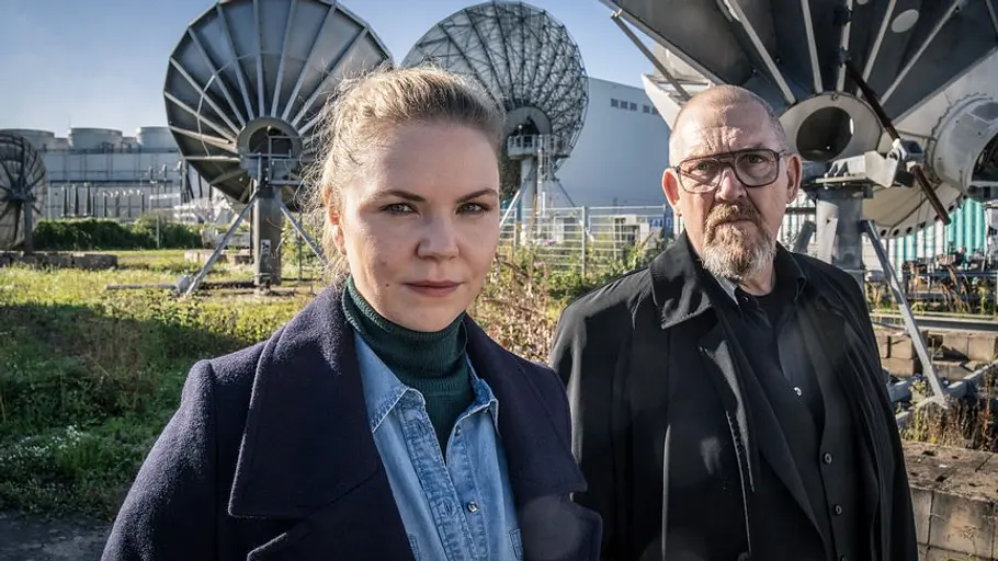 Sie wird die Neue: Ricarda Seifried spielt Kriminalhauptkommissarin Merle Rautenberg. Ricarda Seifried als Kriminalhauptkommissarin Merle Rautenberg im dunklen Mantel und Dietmar Bär mit Brille stehen vor mehreren großen Satellitenschüsseln.