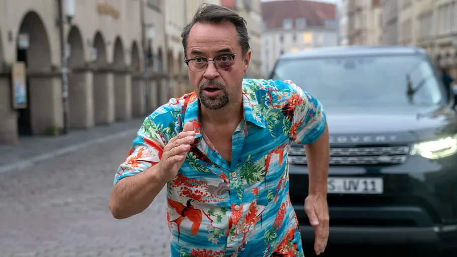 Tatort - Propheteus Jan Josef Liefers läuft in einem Hawaiihemd vor einem Auto weg.