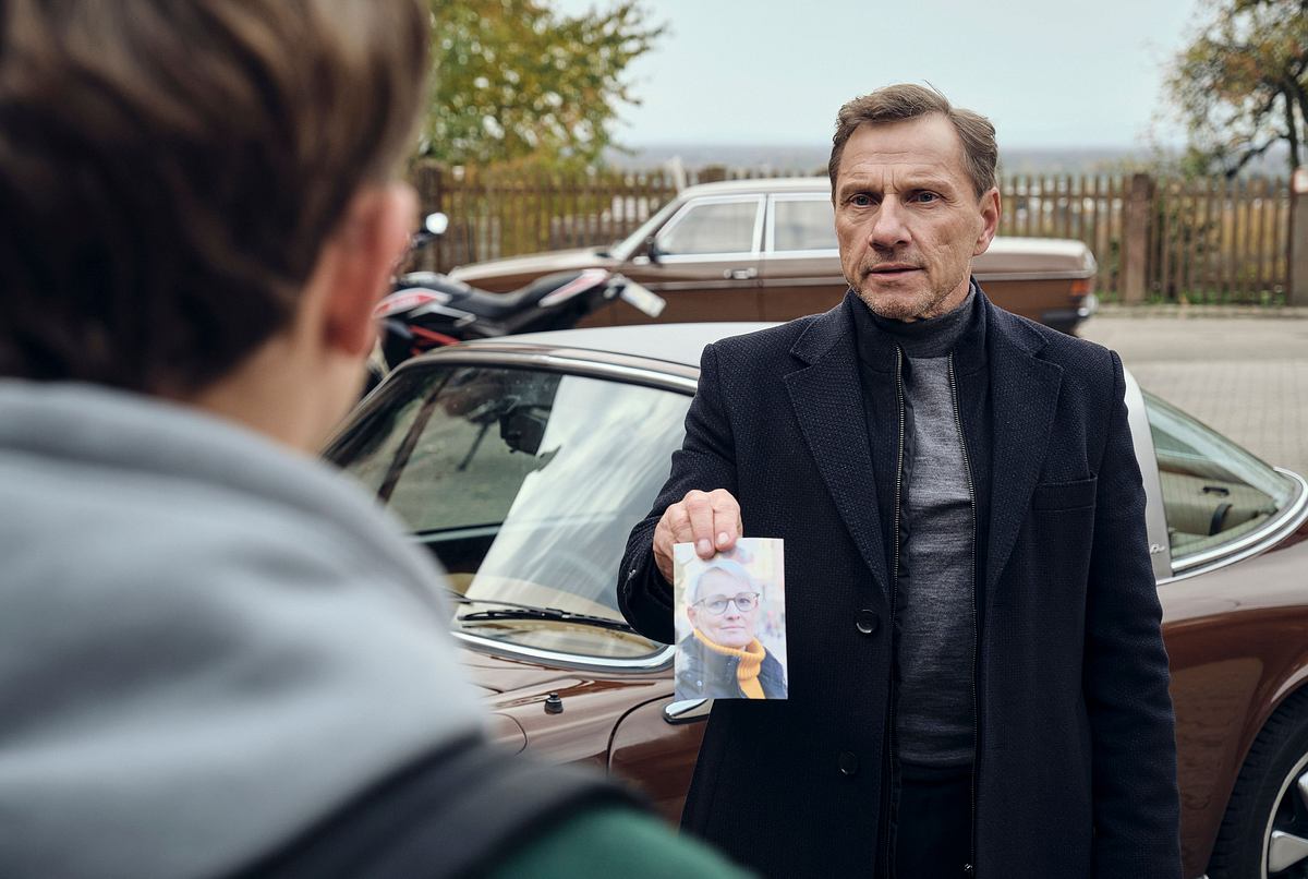 Richy Müller steht vor einem Oldtimer und zeigt Louis Guillaume ein Bild von einer Frau.