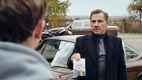 Richy Müller steht vor einem Oldtimer und zeigt Louis Guillaume ein Bild von einer Frau. - Foto: SWR/Benoît Linder