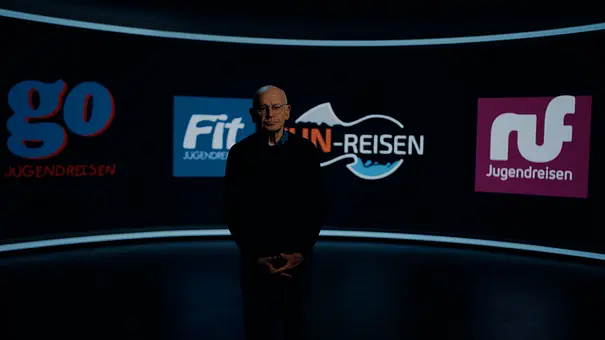 Günther Wallraff steht in einem dunklen Studio vor einer gebogenen Wand mit Logos von Jugendreiseanbietern. - Foto: RTL
