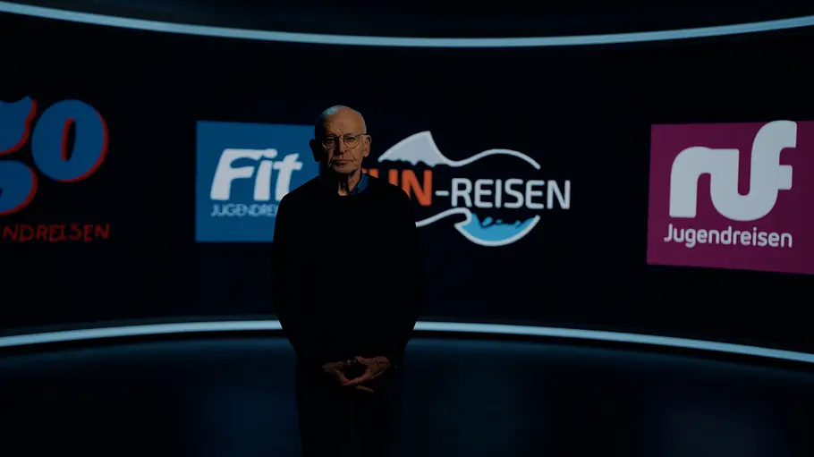 Günther Wallraff Günther Wallraff steht in einem dunklen Studio vor einer gebogenen Wand mit Logos von Jugendreiseanbietern.