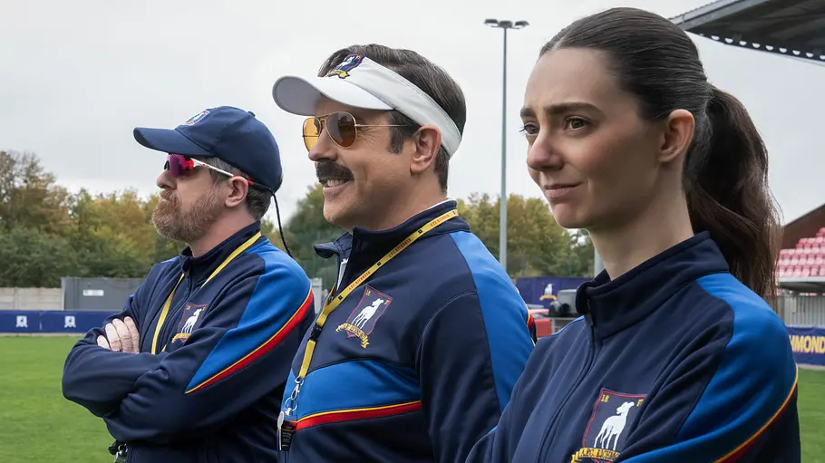 Alice Chilton Ted Lasso Coach Beard, Ted Lasso und Alice stehen von links nach rechts in blau-dunkelblauen Trainingsjacken am Fußballplatz.