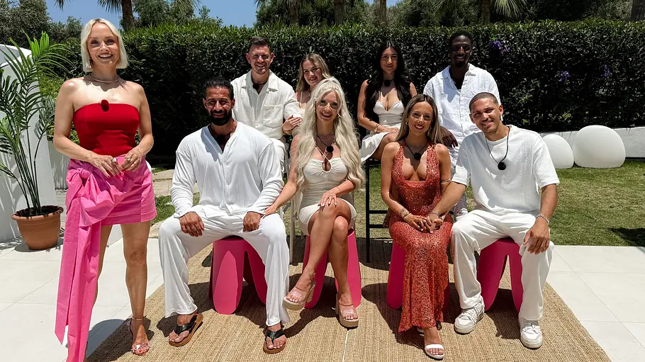 Temptation Island Gruppenfoto von neun Erwachsenen in einer sonnigen Gartenanlage. Die Personen sind auf Stühlen und Hockern arrangiert oder stehen dahinter und blicken lächelnd in die Kamera. Die meisten tragen helle Sommerkleidung in Weiß- und Cremetönen, während eine Frau links im Bild mit einem roten trägerlosen Oberteil und einem pinken Rock auffällt. Mehrere Personen tragen sichtbare Ansteckmikrofone, was auf eine Fernseh- oder Reality-TV-Produktion hindeutet. Im Hintergrund sind Rasen, Hecken, Palmen und weiße Dekorationselemente zu sehen.