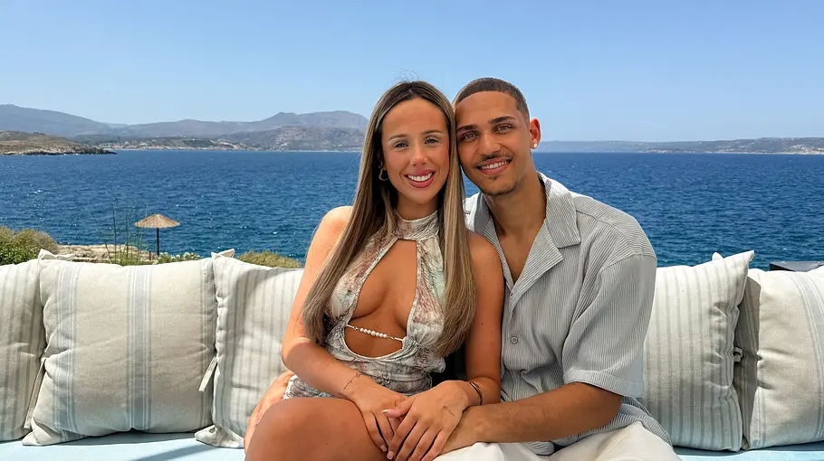 Charleen (21) & Etienne (22) Ein Paar sitzt eng beieinander auf einer Lounge mit Blick auf das Meer. Beide lächeln in die Kamera, während sie sich umarmen. Die Frau trägt ein sommerliches Kleid mit Cut-outs, der Mann ein helles Kurzarmhemd. Im Hintergrund erstrecken sich die blaue Küste, das offene Wasser und eine bergige Landschaft unter klarem Himmel.