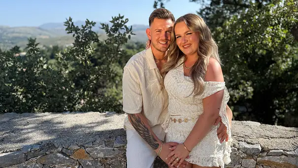 Ein junges Paar steht eng umschlungen vor ihrer Temptation Island-Reise vor einer Steinmauer mit Blick auf eine grüne Hügellandschaft im warmen Abendlicht. - Foto: RTL Ein junges Paar steht eng umschlungen vor ihrer Temptation Island-Reise vor einer Steinmauer mit Blick auf eine grüne Hügellandschaft im warmen Abendlicht. - Foto: RTL