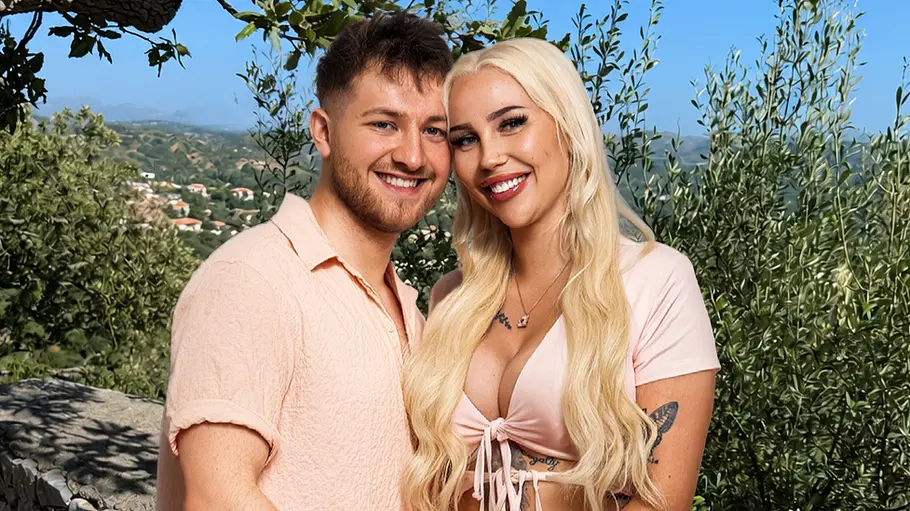 Lisa (24) & Marcel (25) Ein lächelndes Paar posiert eng umschlungen vor einer Steinmauer inmitten einer mediterranen Landschaft. Beide tragen farblich abgestimmte, rosafarbene Sommeroutfits. Im Hintergrund sind Olivenbäume, grüne Hügel und ein wolkenloser Himmel zu sehen. Die Aufnahme vermittelt eine romantische und entspannte Urlaubsstimmung.
