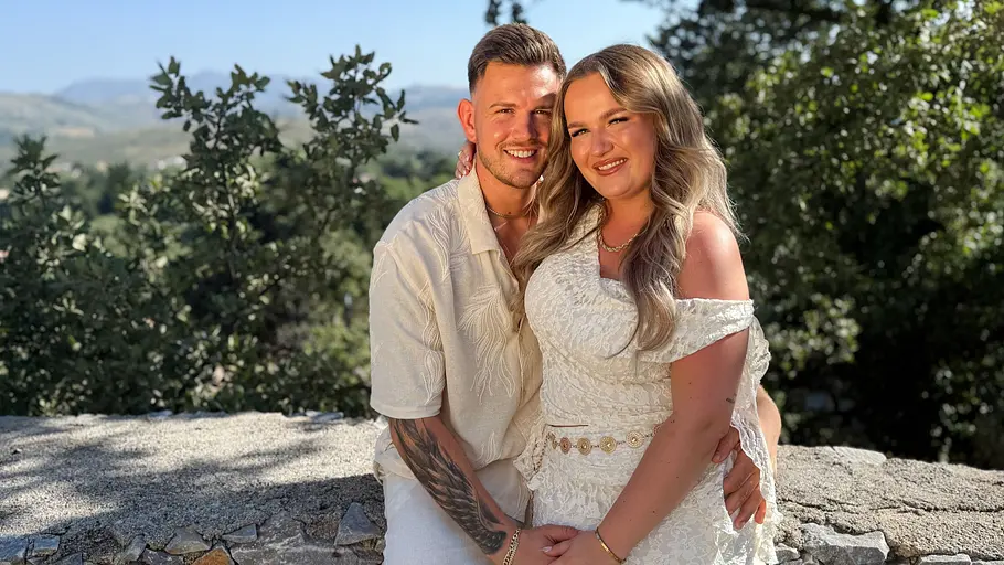 Loucia (23) & Dominik (26) Ein Paar in hellen Sommeroutfits posiert eng umschlungen vor einer Naturkulisse mit Bäumen und Berglandschaft. Die Frau trägt ein weißes Spitzenkleid, der Mann ein helles Hemd und eine weiße Hose. Beide lächeln in die Kamera und stehen vor einer niedrigen Steinmauer im warmen Sonnenlicht.