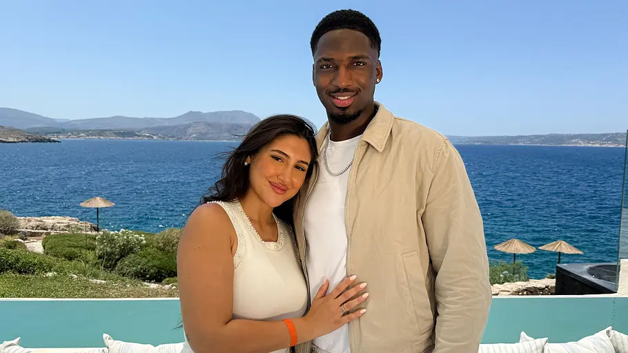 Melina (22) & Jonathan (24) Ein lächelndes Paar posiert auf einer Terrasse mit Blick auf das Meer. Die Frau in einem hellen Kleid lehnt sich an den Mann, der eine beigefarbene Jacke über einem weißen Shirt trägt. Im Hintergrund sind die Küste, das blaue Wasser und Sonnenschirme zu sehen. Die Aufnahme vermittelt eine sommerliche und entspannte Urlaubsatmosphäre.