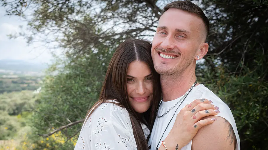 Gloria Schwesinger und Micha Scherer wollen ihre Liebe testen. Gloria mit langen dunklen Haaren und Micha mit kurzen Haaren in weißen Oberteilen umarmen sich lächelnd vor grüner Landschaft.