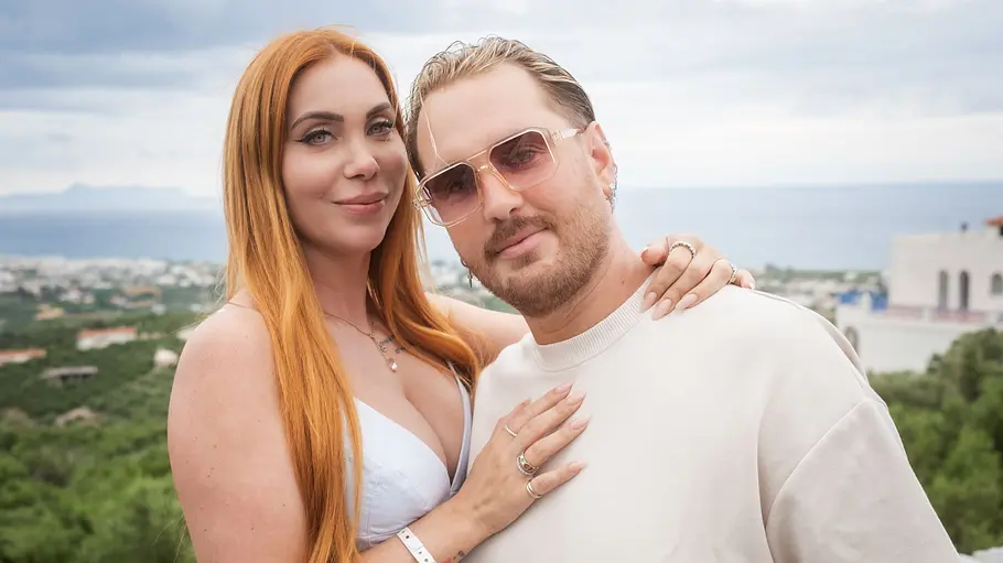 Tara und Dennis Tara und Dennis stehen eng beieinander vor einer grünen Küstenlandschaft. Sie trägt ein helles Kleid, er eine Sonnenbrille.