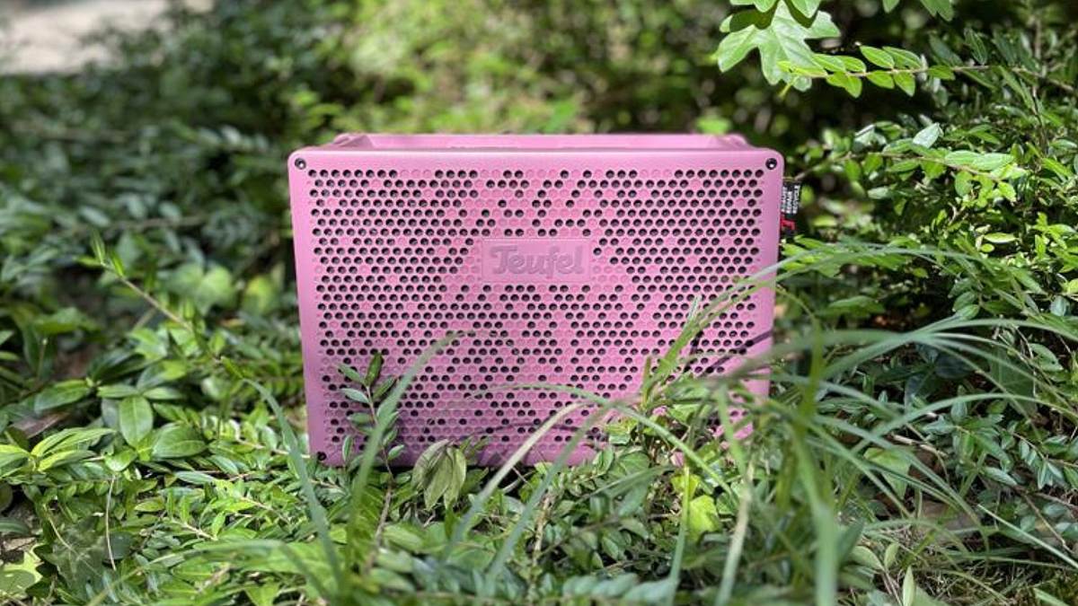 Teufel Bluetooth-Lautsprecher MYND Im Vordergrund sieht man dem Teufel MYND in der Farbe Wild Berry in einer Garten-Umgebung. Im Hintergrund sind grüne Gräser und Sträucher zu sehen.