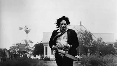 Leatherface rennt mit einer Kettensäge auf einem Feldweg. Hinter ihm ist ein Haus. - Foto: IMAGO / Cinema Publishers Collection Leatherface rennt mit einer Kettensäge auf einem Feldweg. Hinter ihm ist ein Haus. - Foto: IMAGO / Cinema Publishers Collection