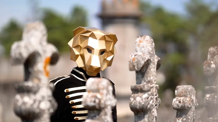 The 50 Der „Löwe“ aus der Show „The 50“ steht mit goldener Maske und schwarzer Uniformjacke auf einem Schlossgelände zwischen steinernen Figuren.
