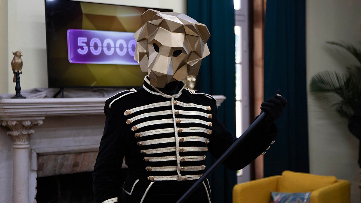 „The 50“-Löwe Der The 50-Löwe steht mit einem Stock in der Hand vor einem Bildschirm auf dem die Zahl 50.000 zu sehen ist