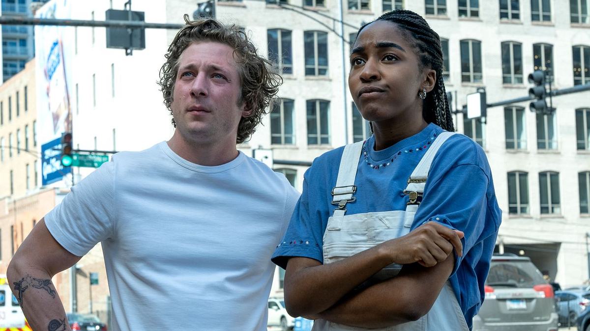 Jeremy Allen White (links) und Ayo Edebiri schauen nach links oben an einer Kreuzung in Chicago. White trägt ein weißes T-Shirt und Edebiri ein blaues T-Shirt mit einer weißen Latzhose. 
