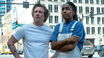 Jeremy Allen White (links) und Ayo Edebiri schauen nach links oben an einer Kreuzung in Chicago. White trägt ein weißes T-Shirt und Edebiri ein blaues T-Shirt mit einer weißen Latzhose.  - Foto: Hulu / Disney+
