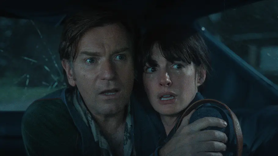 Anne Hathaway und Ewan McGregor schauen schockiert aus einem beschädigten Auto.