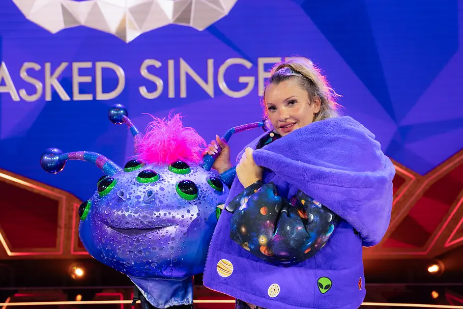 Evelyn Burdecki steckte unter Cosma Evelyn Burdecki steht im violetten Kostüm neben einem großen, glitzernden Alienkopf mit mehreren Augen und pinken Haaren.