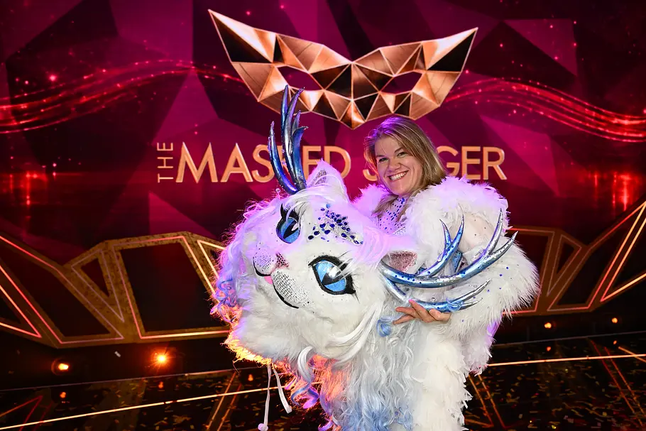 Gisa Flake überraschte als Yeti Cat Gisa Flake hält im weißen Fellkostüm einen großen Fantasietierkopf mit blauem Geweih vor dem Logo von „The Masked Singer“.
