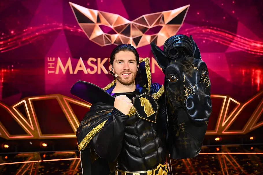 Joey Heindle steckte unter P.S. Joey Heindle posiert im schwarz-goldenen Kostüm mit erhobener Faust neben einer Pferdemaske vor dem „The Masked Singer“-Logo.