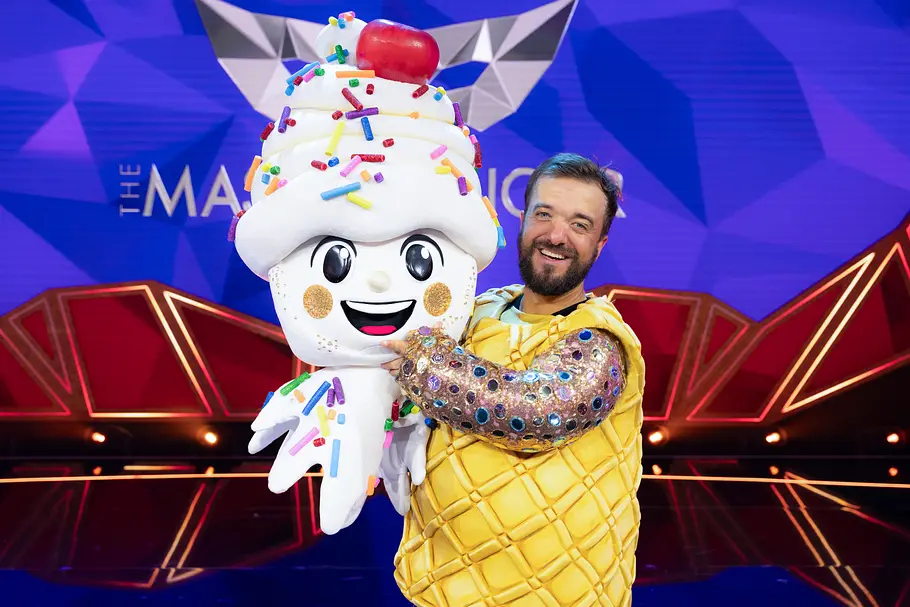 Mathias Mester war Luigi Eishörnchen Mathias Mester trägt ein gelbes Waffelkostüm mit Glitzerärmeln und hält einen großen Softeis-Kostümkopf mit bunten Streuseln.