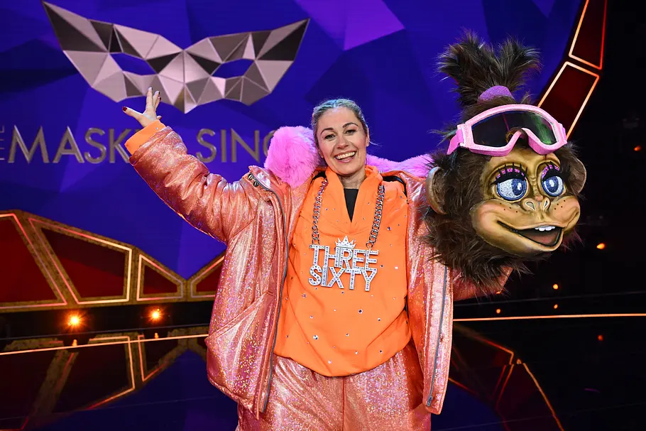 Ruth Moschner wechselte als Three Sixty die Seiten Ruth Moschner lächelt im orange-rosa Glitzeroutfit mit erhobenem Arm neben einem großen Affenkostümkopf mit rosa Skibrille.