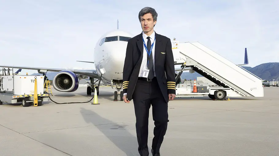 The Rehearsal Nathan Fielder läuft in Pilotenuniform über das Rollfeld vor einem Passagierflugzeug. Im Hintergrund sind eine Fluggasttreppe und Bodenfahrzeuge zu sehen – Szene aus der HBO-Serie „The Rehearsal“.