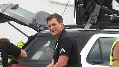 Nathan Fillion steht lächelnd in schwarzer Polizeiuniform vor einem schwarz-weißen Fahrzeug mit Ausrüstung auf dem Dach. - Foto: IMAGO / Avalon.red