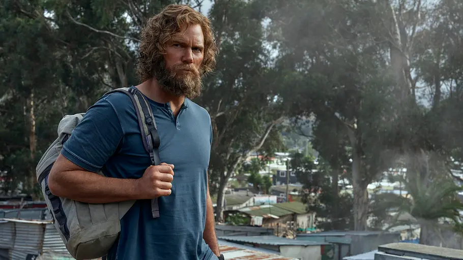 „The Terminal List“ Staffel 2: Reece lebt im Versteck James Reece steht mit Bart, blauem Shirt und grauem Rucksack vor Wellblechhütten und Bäumen.