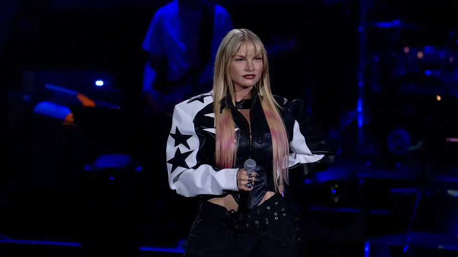 The Voice Folge 3 Emma Powda Emma Powda steht selbstbewusst auf der Bühne von The Voice. Sie trägt eine schwarz-weiße Lederjacke mit Sternen. und hat rosa gefärbte Strähnen im Haar.
