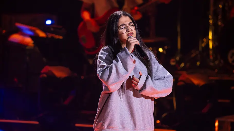 The Voice Folge 5: Aylin Basar Aylin Basar singt mit Brille und grauem Sweatshirt in ein Mikrofon. Hinter ihr sind Musiker auf der dunklen Bühne zu sehen.