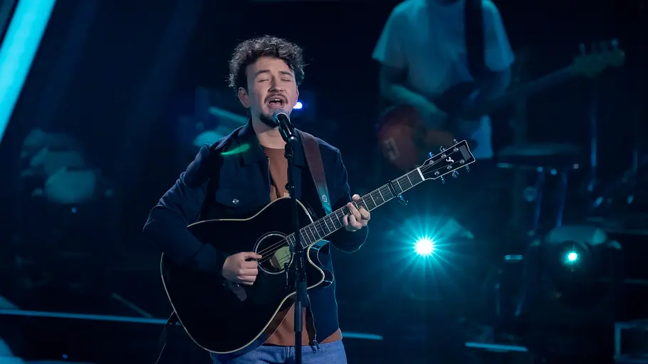 The Voice: Folge 7 Bjorn Shabanaga Bjorn Shabanaga singt auf der blau beleuchteten The Voice-Bühne ins Mikrofon und spielt eine schwarze Akustikgitarre.