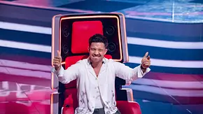 Nico Santos sitzt im weißen Hemd auf einem roten „The Voice“-Sessel und streckt lächelnd beide Daumen nach oben. - Foto: Joyn Nico Santos sitzt im weißen Hemd auf einem roten „The Voice“-Sessel und streckt lächelnd beide Daumen nach oben. - Foto: Joyn