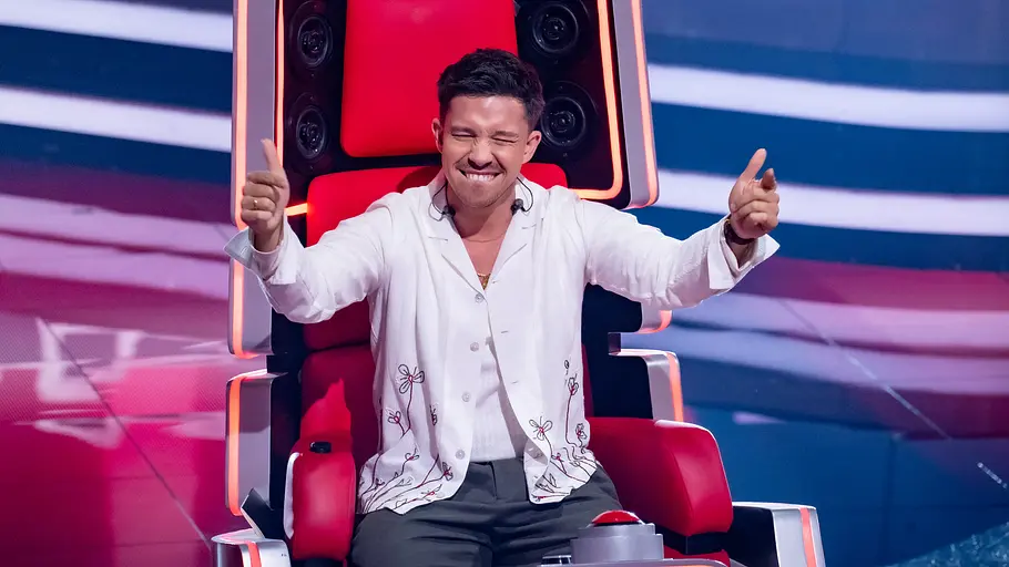 The Voice Folge 7: Nico Santos Nico Santos sitzt im weißen Hemd auf einem roten „The Voice“-Sessel und streckt lächelnd beide Daumen nach oben.