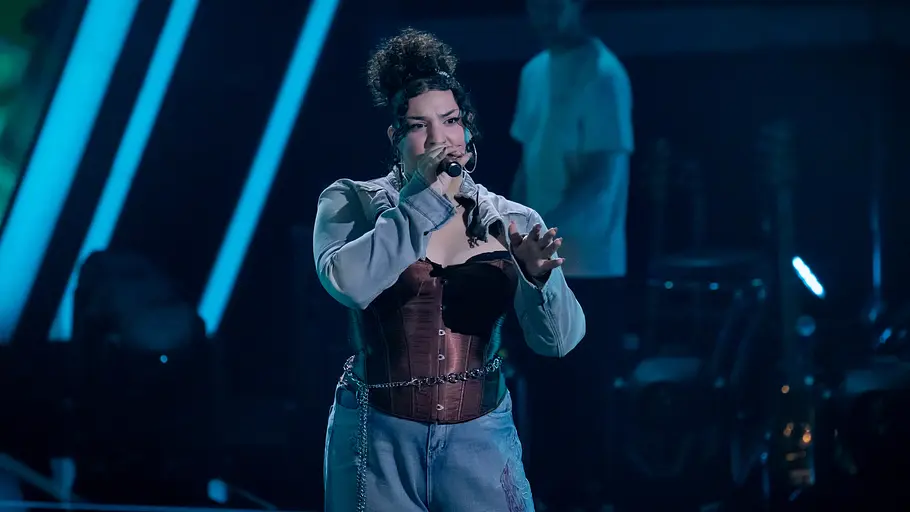the Voice: Folge 8 Mel Vardouniotis Mel Vardouniotis singt mit einem Mikrofon auf einer blau beleuchteten Bühne. Sie trägt ein braunes Korsett und helle Jeans.