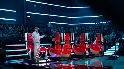 Rea Garvey steht mit ausgebreiteten Armen neben roten Jurystühlen bei „The Voice“. Er ist als einziger zur Bühne gedreht. Dahinter sitzt Publikum im blauen Licht. - Foto: Joyn