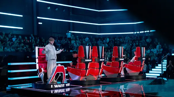 Rea Garvey steht mit ausgebreiteten Armen neben roten Jurystühlen bei „The Voice“. Er ist als einziger zur Bühne gedreht. Dahinter sitzt Publikum im blauen Licht. - Foto: Joyn Rea Garvey steht mit ausgebreiteten Armen neben roten Jurystühlen bei „The Voice“. Er ist als einziger zur Bühne gedreht. Dahinter sitzt Publikum im blauen Licht. - Foto: Joyn
