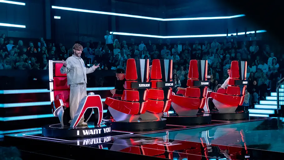 The Voice Folge 8: Rea Garvey Rea Garvey steht mit ausgebreiteten Armen neben roten Jurystühlen bei „The Voice“. Er ist als einziger zur Bühne gedreht. Dahinter sitzt Publikum im blauen Licht.