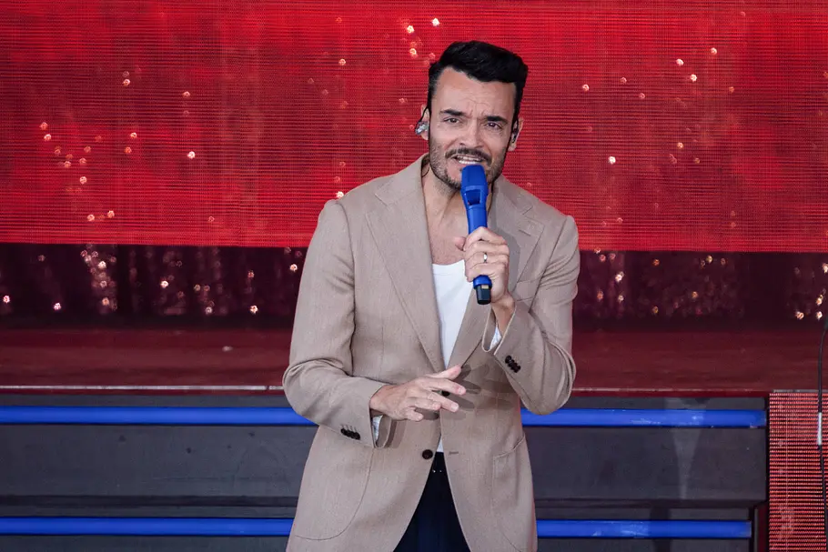 „The Voice“ Gast-Coach Giovanni Zarrella. Giovanni Zarrella im beigen Sakko hält ein blaues Mikrofon vor den Mund und gestikuliert auf einer Bühne mit rotem Hintergrund.